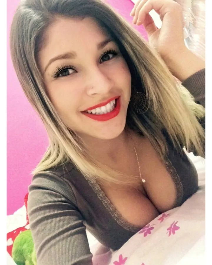 Jeune femme blonde, portant un haut blanc et un short rose, prenant un selfie dans un miroir, cheveux longs, lunettes.